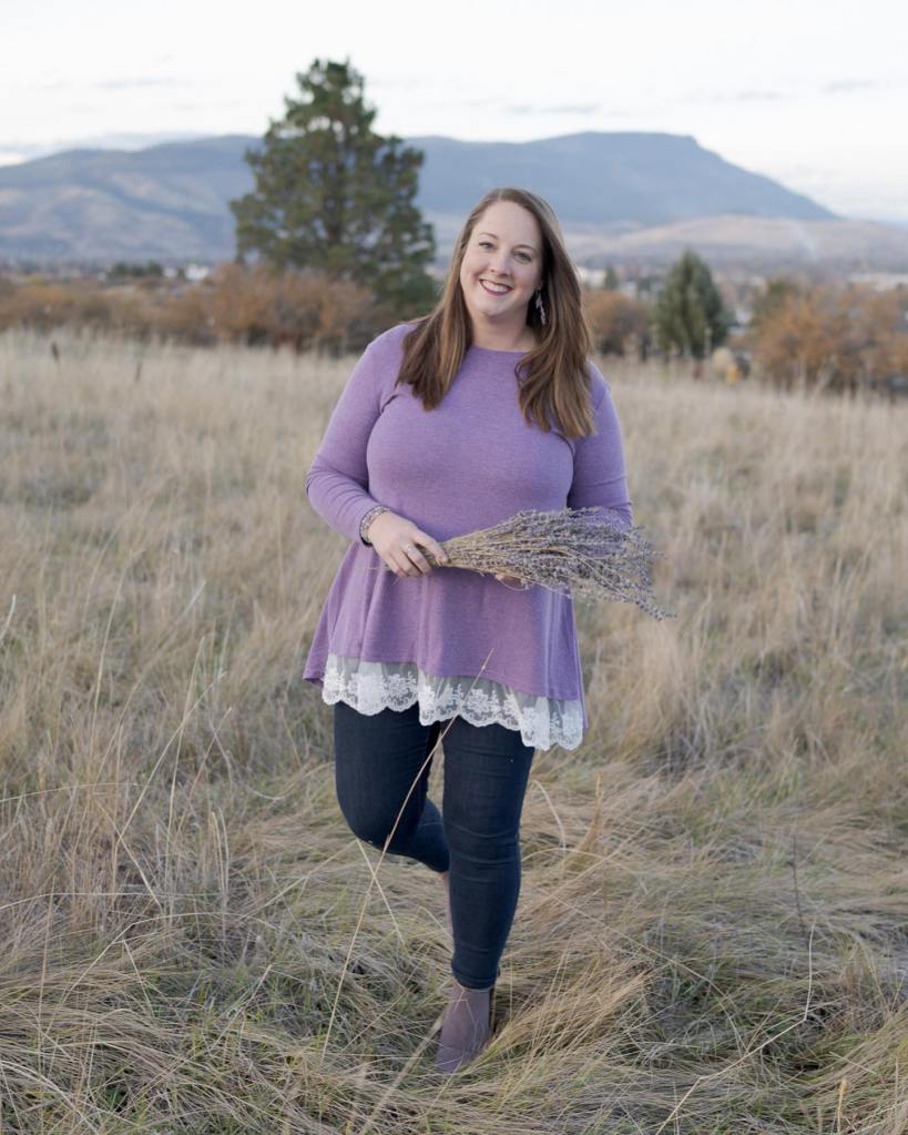A smiling woman holding a bundle of lavender stands in a grassy field with mountains in the background, wearing a purple top and dark jeans.