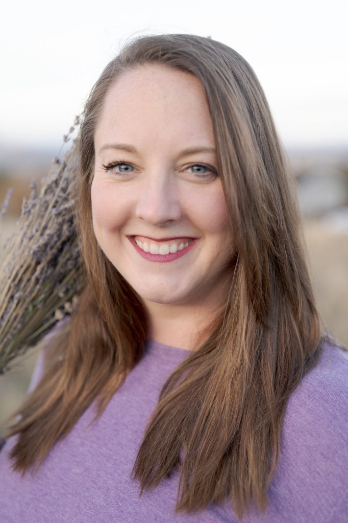 A woman with long brown hair smiling, wearing a purple shirt, and holding a bundle of lavender against a blurred outdoor background.