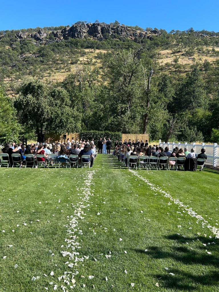 A picturesque outdoor wedding ceremony scene with guests seated on chairs arranged on a lawn, flower petals scattered along the aisle, and a backdrop of trees and rocky hills.