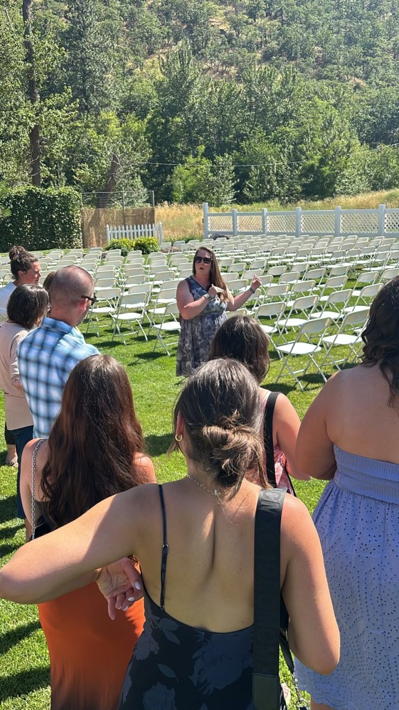 A group of people gathered outdoors in a grassy area, listening attentively to a woman speaking, with rows of empty chairs set up for a wedding ceremony in the background.
